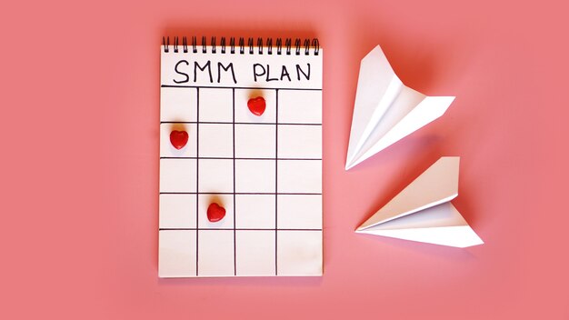 Social-Media-Marketingkonzept - SMM-Plan in einem Notizbuch auf rosafarbenem Hintergrund mit Süßigkeiten in Form von Herzen und Papierflugzeugen