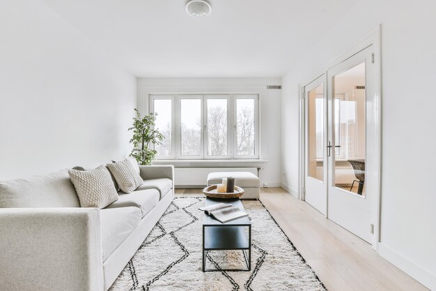 Sofá confortável com almofadas localizado no carpete perto da mesa em uma sala bem iluminada com janela e porta em um apartamento moderno