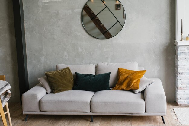 Sofá gris con almohadas espejo redondo colgado en la pared en una moderna sala de estar de estilo escandinavo