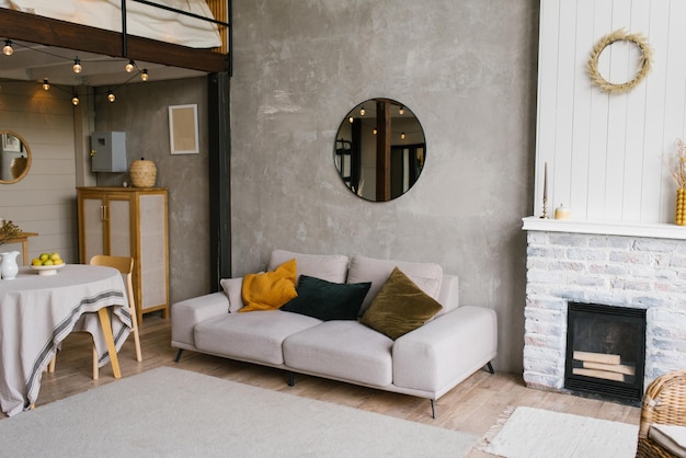 Sofá gris con almohadas, un espejo redondo cuelga de la pared en una moderna sala de estar de estilo escandinavo cerca de la chimenea y la mesa de comedor