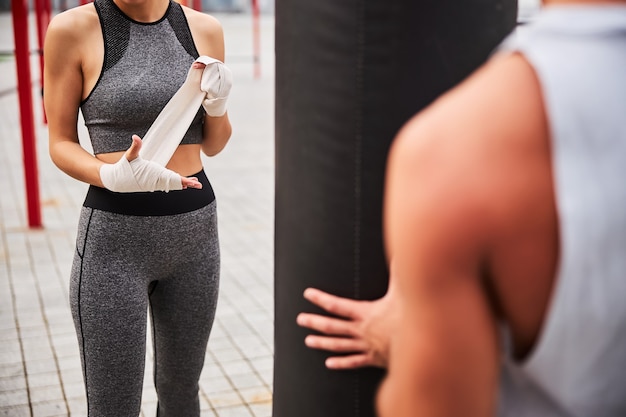 El sofá masculino está ayudando a la mujer delgada con vendas en las manos durante el entrenamiento al aire libre