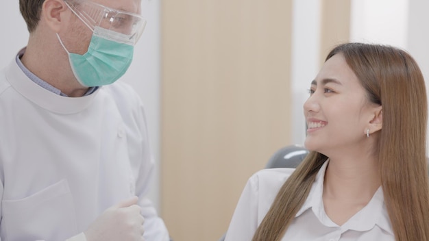 En un sofisticado centro dental, un dentista caucásico con mascarilla explica cómo tratar los dientes a una alegre paciente asiática