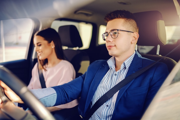 Sofisticado jovem casal caucasiano feliz dirigindo carro. homem de mãos dadas no volante.