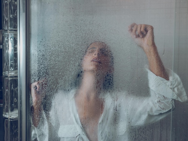 Sofrendo mulher molhada de camisa branca fechando os olhos e levantando os braços enquanto estava atrás da cortina de polietileno na cabine do chuveiro