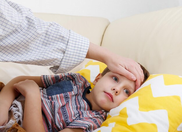 Sohn wird krank und seine Mutter überprüft die Temperatur. Kranker Junge liegt im Bett und die Hand der Mutter misst die Temperatur.