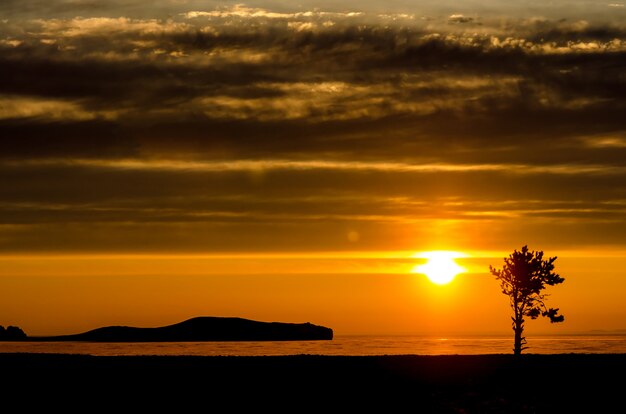 Sol acima do lago, montanha e árvore ao pôr do sol na luz de contraste com nuvens