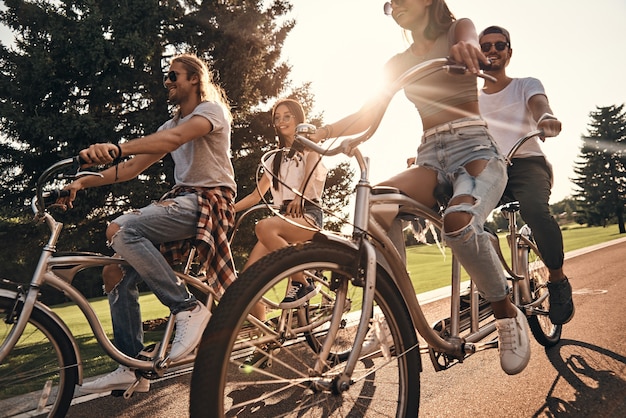 Foto sol cálido y gran compañía. grupo de jóvenes felices en ropa casual sonriendo mientras andan en bicicleta juntos al aire libre