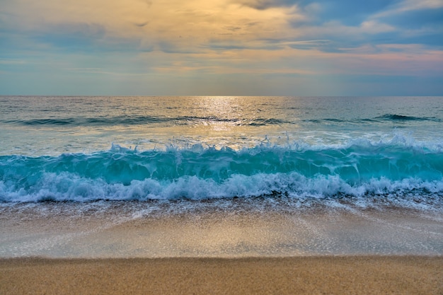 El sol detrás de las nubes se refleja en el agua y las olas con espuma golpean la arena.