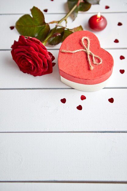 Una sola flor de rosa roja fresca sobre la mesa de madera blanca con lentejuelas en forma de corazón esparcidas alrededor de velas y regalo en caja San Valentín o concepto de amor con espacio para copiar