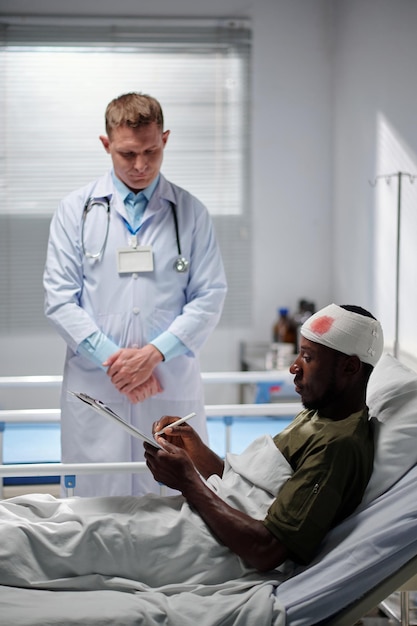 Soldado afro-americano deitado na cama na enfermaria e assinando documento fornecido pelo médico