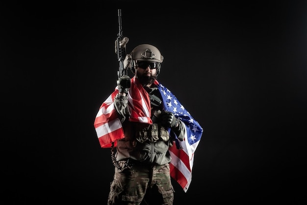Soldado americano em uniforme militar com uma arma segura a bandeira dos EUA contra um fundo escuro