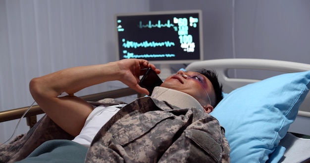 Soldado asiático ferido com colarinho no pescoço deitado na cama do hospital e falando no smartphone
