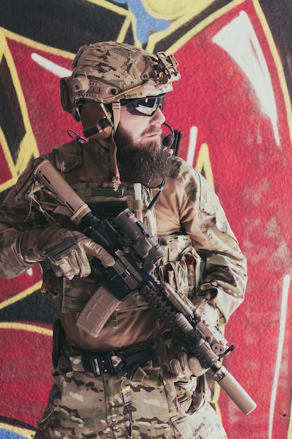 Un soldado barbudo en un uniforme de las fuerzas especiales se encuentra frente a una pared dibujada con un rifle en la mano enfoque selectivo