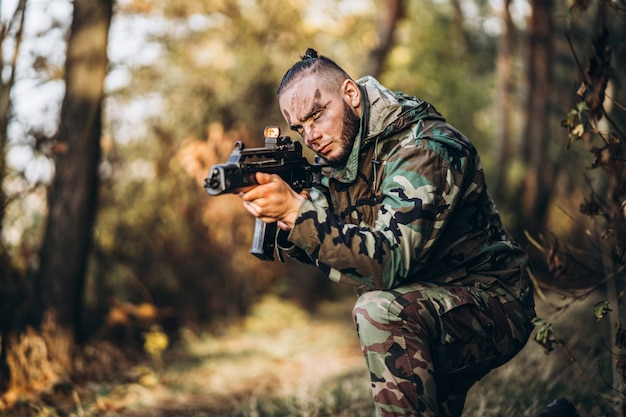 Soldado camuflado com rifle e rosto pintado, jogando airsoft ao ar livre na floresta