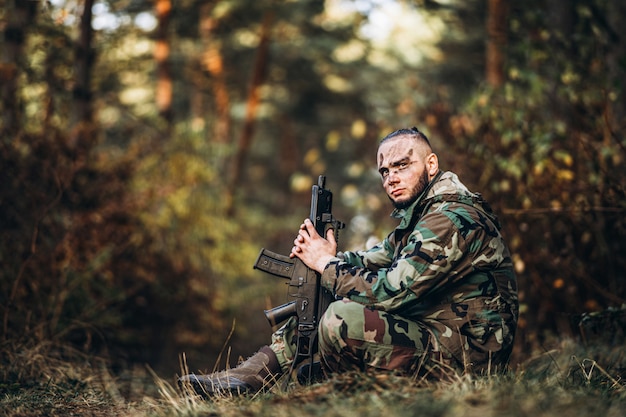 Soldado camuflado com rifle e rosto pintado, sentado na grama.