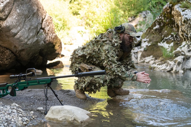 Soldado com uniforme camuflado bebendo água doce do rio. Rifle de atirador militar ao lado.