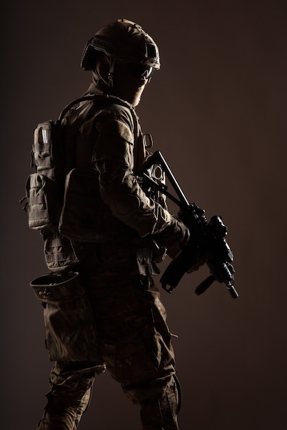 Soldado del ejército con casco, uniforme de batalla con munición táctica, rostro oculto detrás de máscara y gafas, mirando hacia atrás por encima del hombro, escabulléndose en la oscuridad con rifle de servicio en mano, bajo perfil, sesión de estudio