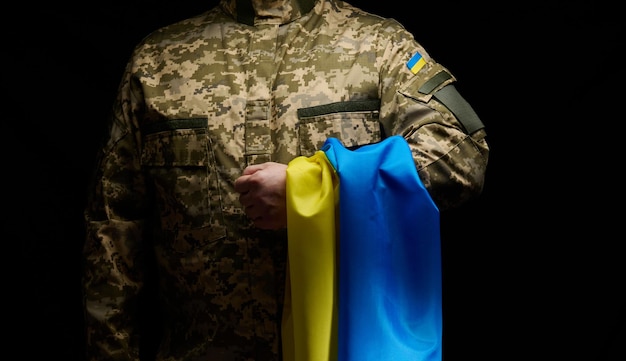 Foto un soldado de las fuerzas armadas ucranianas está de pie con una bandera azul-amarilla de ucrania sobre un fondo negro. honrar a los veteranos y conmemorar a los muertos en la guerra.