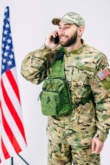 Soldado guapo con gorra y mochila hablando con su familia usando un teléfono móvil después de un entrenamiento militar cerca de la bandera estadounidense en un estudio de fondo blanco