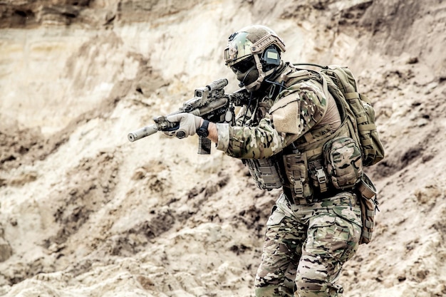 Soldado de infantería del ejército, soldado de las fuerzas especiales con rostro oculto, casco opscore protegido y armadura corporal, auriculares de radio tácticos equipados, con uniforme de camuflaje ocp, apuntando con carabina de servicio en las manos