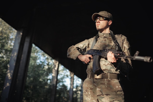 Un soldado masculino serio con un uniforme de camuflaje gris con gafas tácticas con lentes amarillas y una gorra con una ametralladora negra en las manos dentro de una gran fábrica autorizada