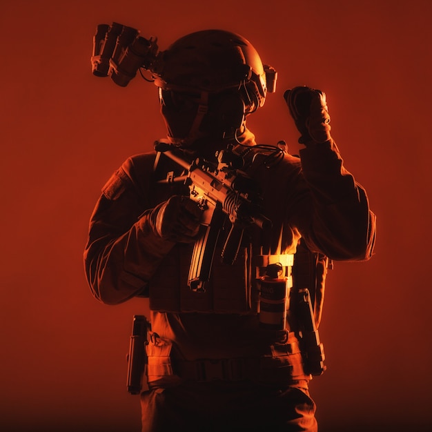 Foto soldado de operaciones especiales, combatiente del equipo swat con máscara y gafas, dispositivo de visión nocturna equipado, rifle armado de servicio de cañón corto, mirando hacia atrás y mostrando la señal de congelación, sesión de estudio de bajo perfil