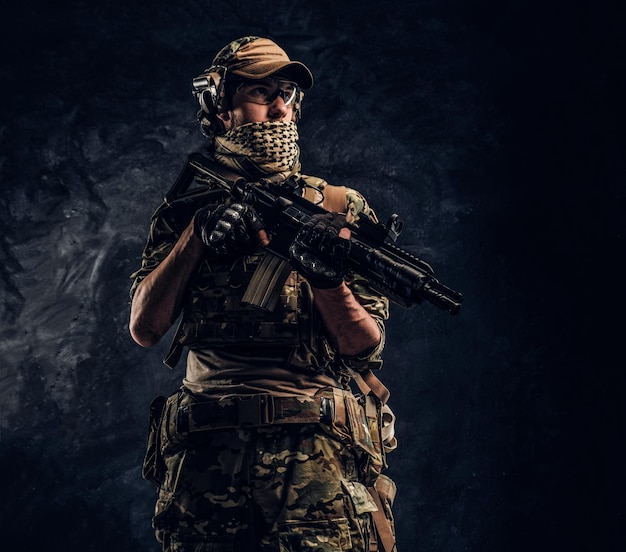 Soldado totalmente equipado em uniforme de camuflagem segurando um rifle de assalto. Foto de estúdio contra uma parede texturizada escura