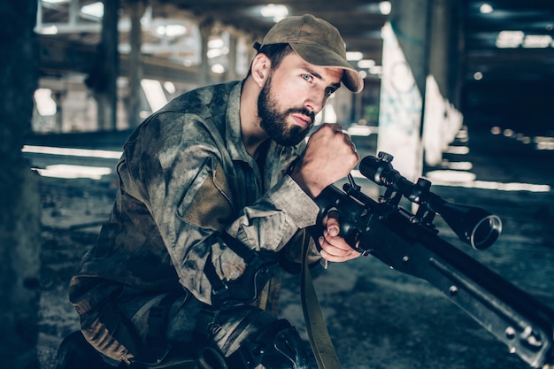 Foto el soldado tranquilo y pacífico mira hacia adelante. el esta esperando. joven está sentado una rodilla. guy usa uniforme militar.