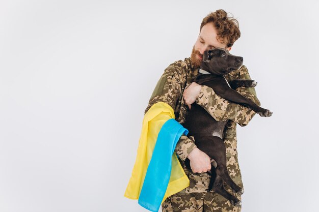 Soldado ucraniano en uniforme militar con una bandera amarilla y azul sostiene un perro en sus brazos sobre un fondo blanco.