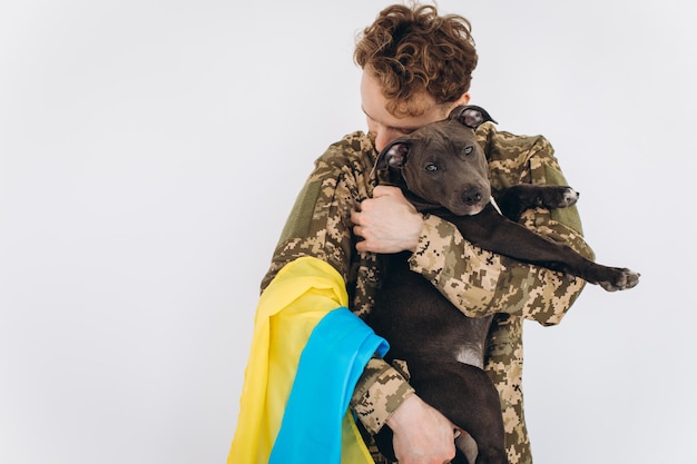 Soldado ucraniano en uniforme militar con una bandera amarilla y azul sostiene un perro en sus brazos sobre un fondo blanco.