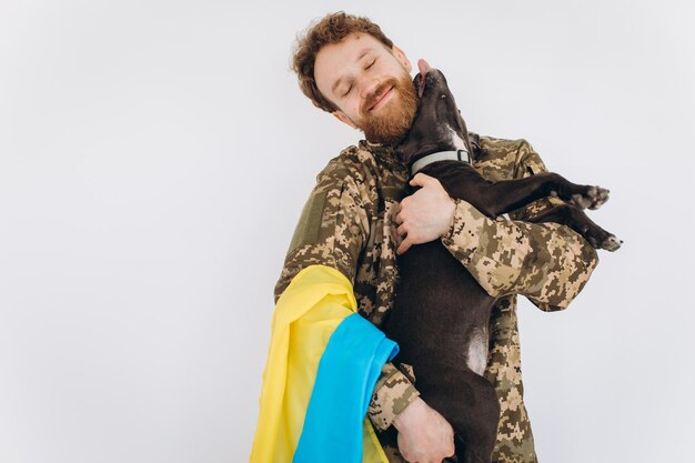 Soldado ucraniano en uniforme militar con una bandera amarilla y azul sostiene un perro en sus brazos sobre un fondo blanco.