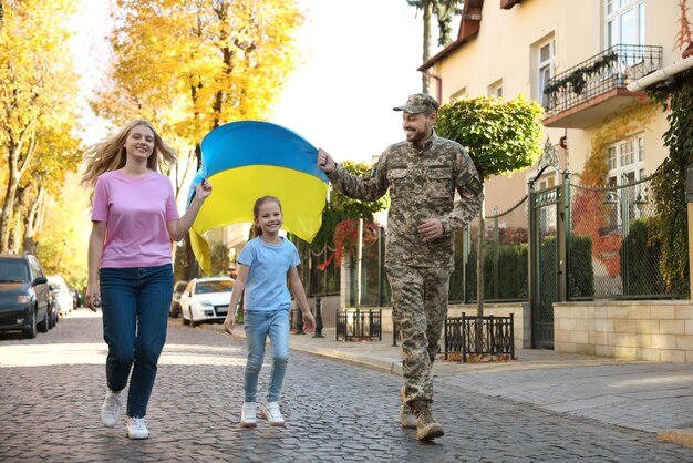 Soldado en uniforme militar con su familia corriendo y sosteniendo la bandera ucraniana en la calle de la ciudad