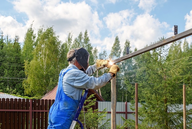 Soldador de homem em uma máscara de soldagem, uniforme de construção e luvas de proteção cozinha metal em um canteiro de obras de rua. Construção de um pavilhão, pérgula perto de uma casa de campo num dia de verão.