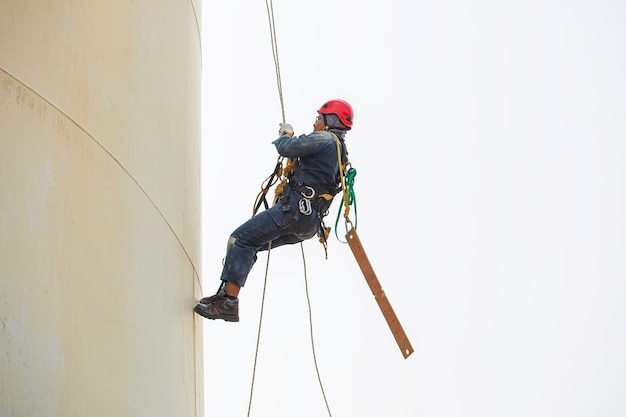 Soldador industrial de acceso por cuerda de trabajador masculino que trabaja en altura usando arnés, inspección de acceso por cuerda de equipo de seguridad de casco de la industria de tanques de almacenamiento de espesor.