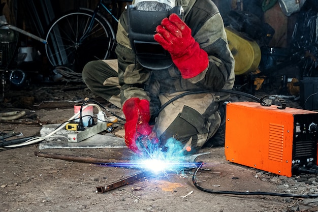 Un soldador con una máscara de soldadura trabaja con un electrodo de arco en su garaje. Soldadura, construcción, trabajos en metal.