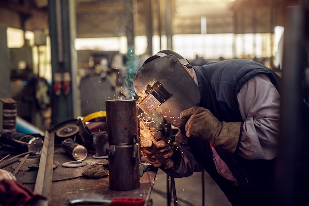 Soldador profissional em uniforme de proteção e máscara de solda de tubo metálico na mesa industrial com outras ferramentas na oficina industrial.