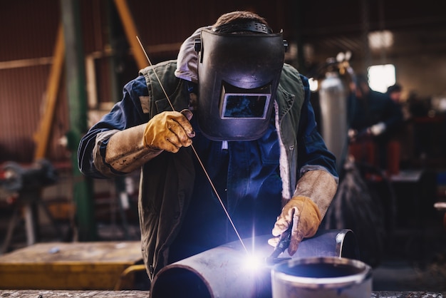 Soldador en uniforme protector y máscara de soldadura de tubo de metal en la mesa industrial mientras vuelan chispas.