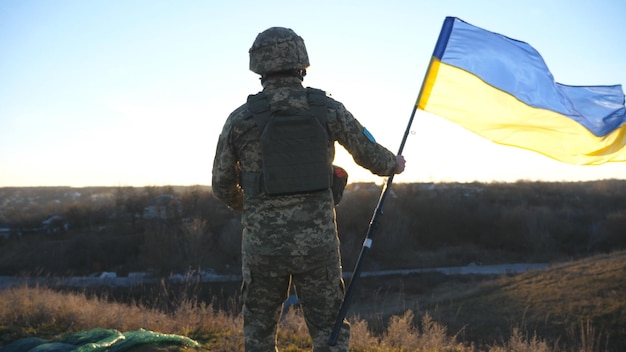 Foto soldat der ukrainischen armee rennt auf den hügel, um die flagge der ukraine zu schwenken. ein mann in militäruniform hob die flagge gegen sonnenuntergang als zeichen des sieges gegen die russische aggression. invasionswiderstand