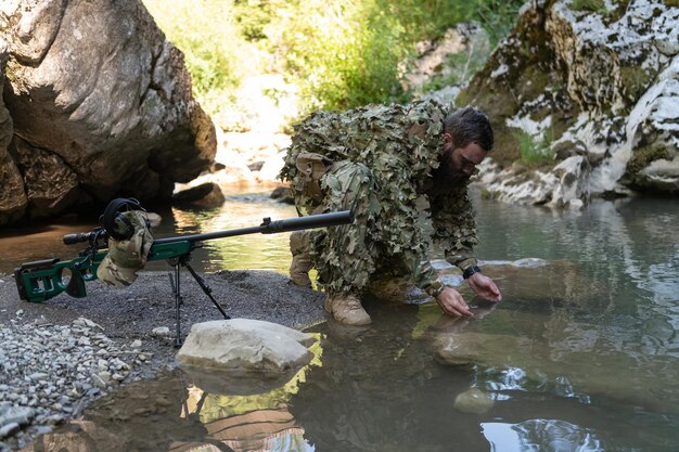 Foto soldat in tarnanzuguniform trinkt frisches wasser aus dem fluss. militärisches scharfschützengewehr an der seite.