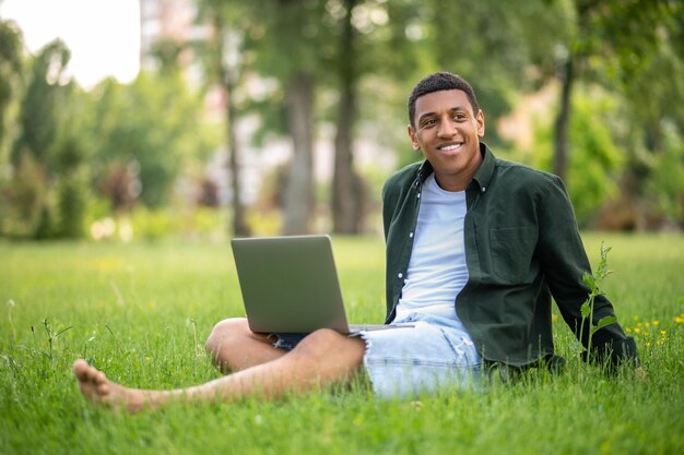 Solidão. Cara sorridente de pele escura descalço com laptop sentado na grama no parque olhando pensativamente para o lado