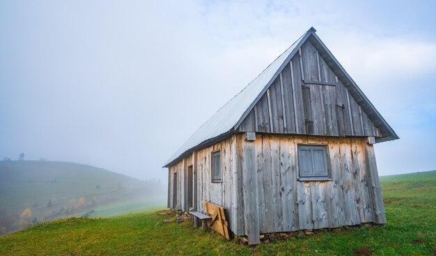 Una solitaria casita gris se encuentra en un prado verde húmedo fresco en medio de una densa niebla gris