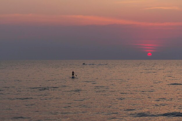 Foto solitario sup board man surfer navega moviéndose en el agua del mar hacia la puesta del sol aventuras en el mar