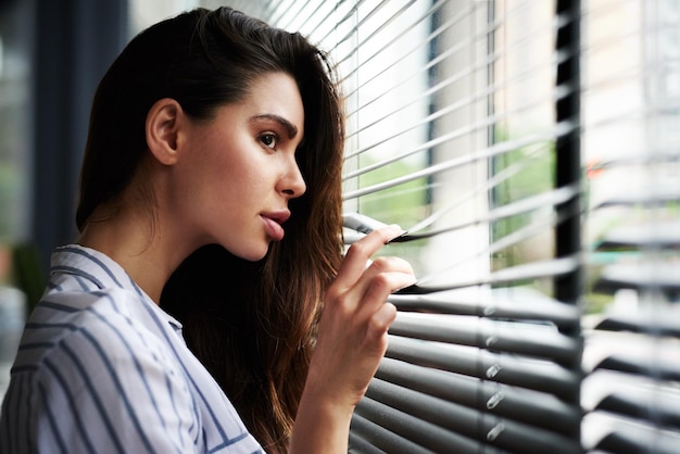 Soll ich heute ausgehen Schnappschuss einer attraktiven jungen Frau, die durch ihr Fenster schaut, während sie zu Hause drinnen steht