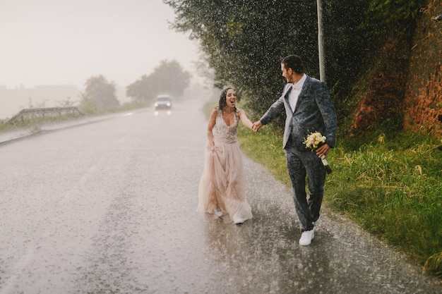 Solo casados cogidos de la mano y caminando sobre la lluvia. Caminando con ropa ceremonial mojada en la carretera. Sonriendo y divirtiéndose.