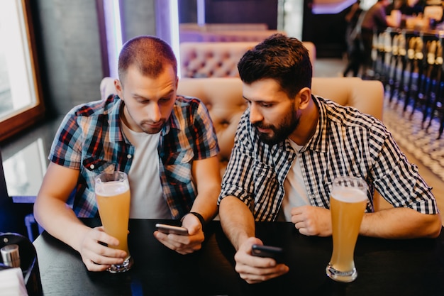 Solo mira esta foto. Dos hombres jóvenes felices en ropa casual bebiendo cerveza en el pub mientras uno de ellos sostiene el teléfono inteligente y lo señala con una sonrisa