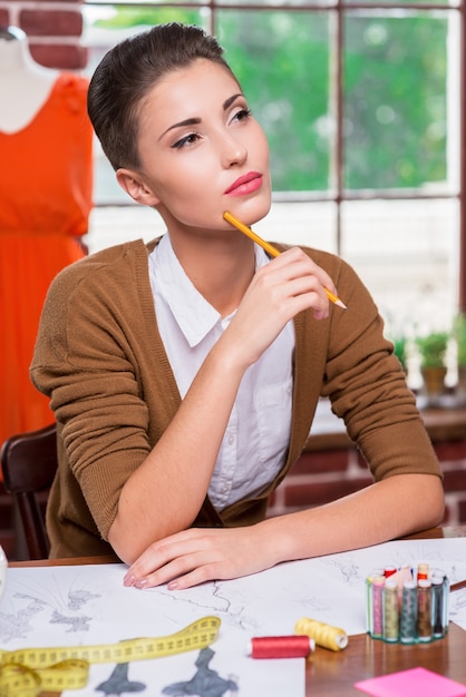 Solo pensamiento creativo. Hermosa joven diseñadora de moda sosteniendo un lápiz y sonriendo a la cámara mientras está sentada en su lugar de trabajo