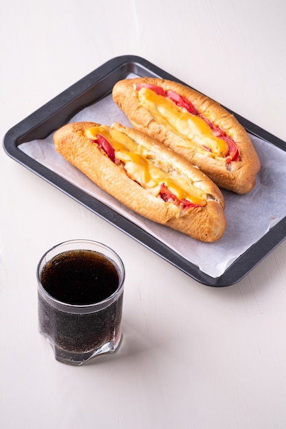 Solo vaso de refresco de cola con dos hot dog comida rápida chatarra fresca salsa de queso salchicha aislado en la bandeja para hornear pared blanca