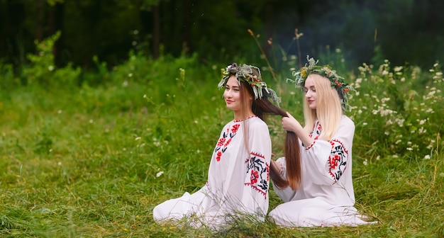 Solstício de verão. duas meninas com roupas eslavas tecem tranças nos cabelos perto do fogo.