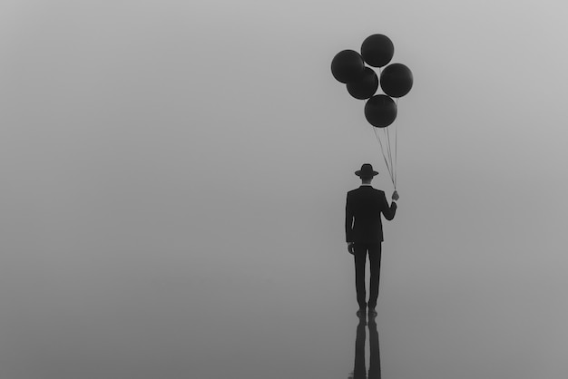 Soltero surrealista en un traje con un sombrero con globos en la mano en el agua por la mañana en la niebla