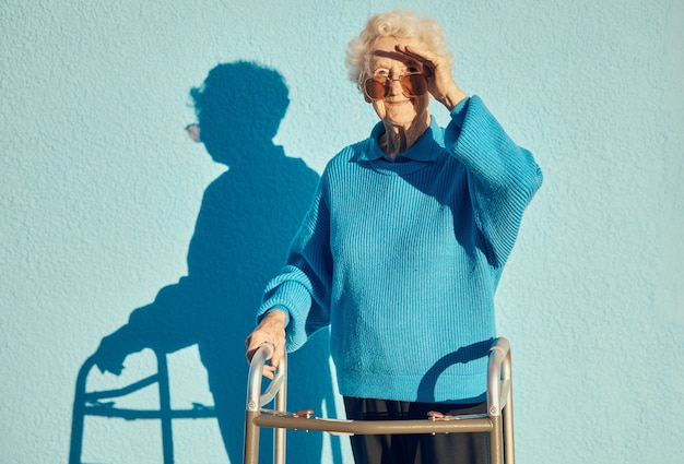 Sombra de retrato y discapacidad con una mujer mayor sobre un fondo de pared azul mientras sostiene un andador de movilidad al aire libre Discapacidad de salud y moda con una mujer madura parada afuera sola